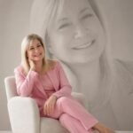 Smiling woman in a pink pantsuit sits in a light chair, with a large grayscale portrait of her on the wall behind her.
