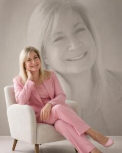 Smiling woman in a pink pantsuit sits in a light chair, with a large grayscale portrait of her on the wall behind her.